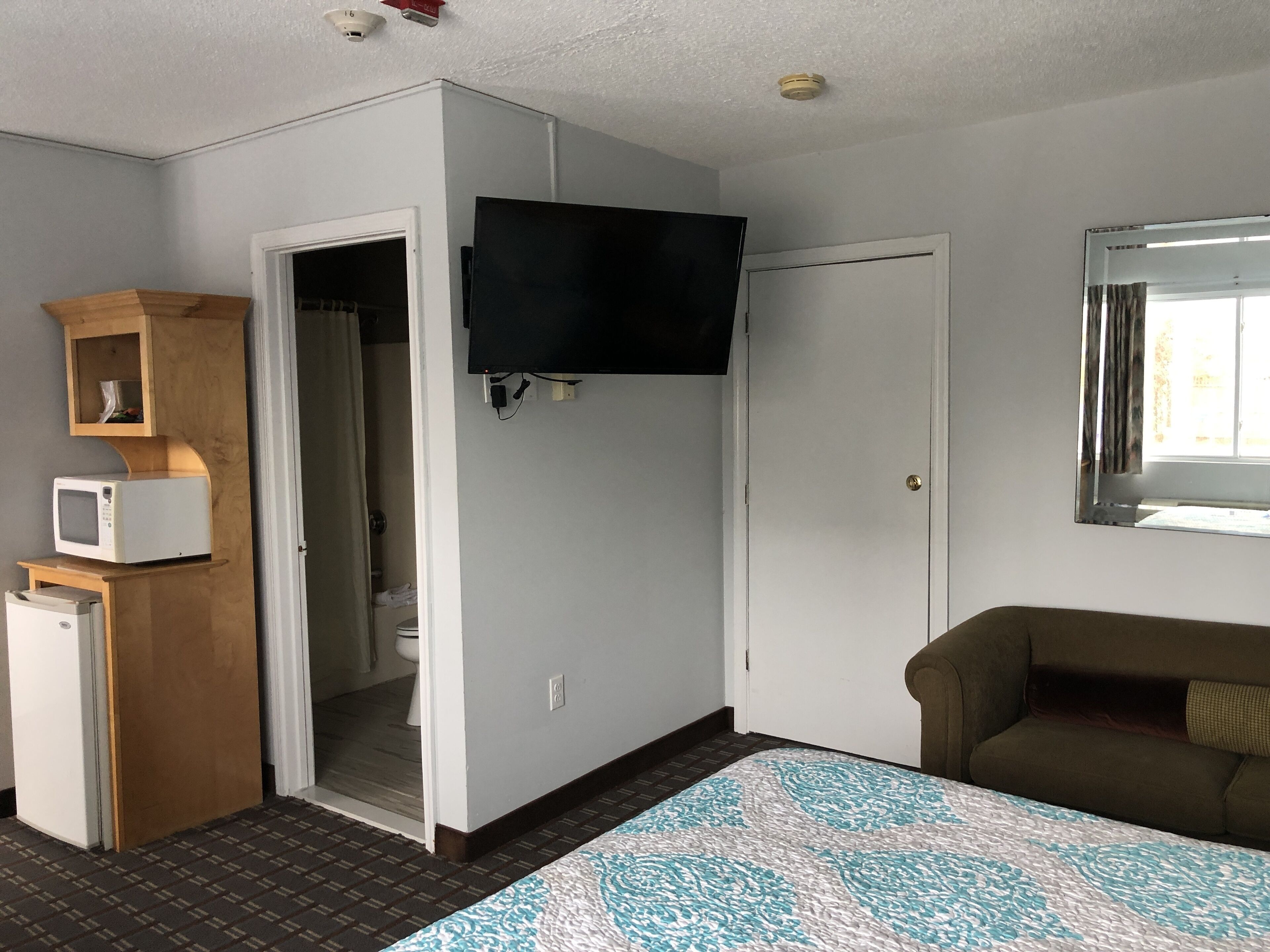 A compact room with a bed, a television mounted on the wall, a microwave, and a small cabinet.