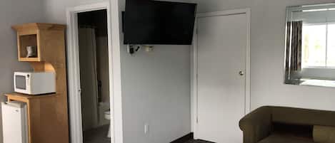 A compact room with a bed, a television mounted on the wall, a microwave, and a small cabinet.