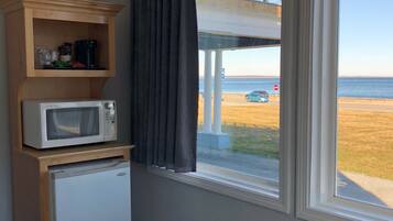 Una habitación con un estante de madera que contiene un microondas y un refrigerador, una ventana con vista a la playa y un muelle, y un aire acondicionado blanco.