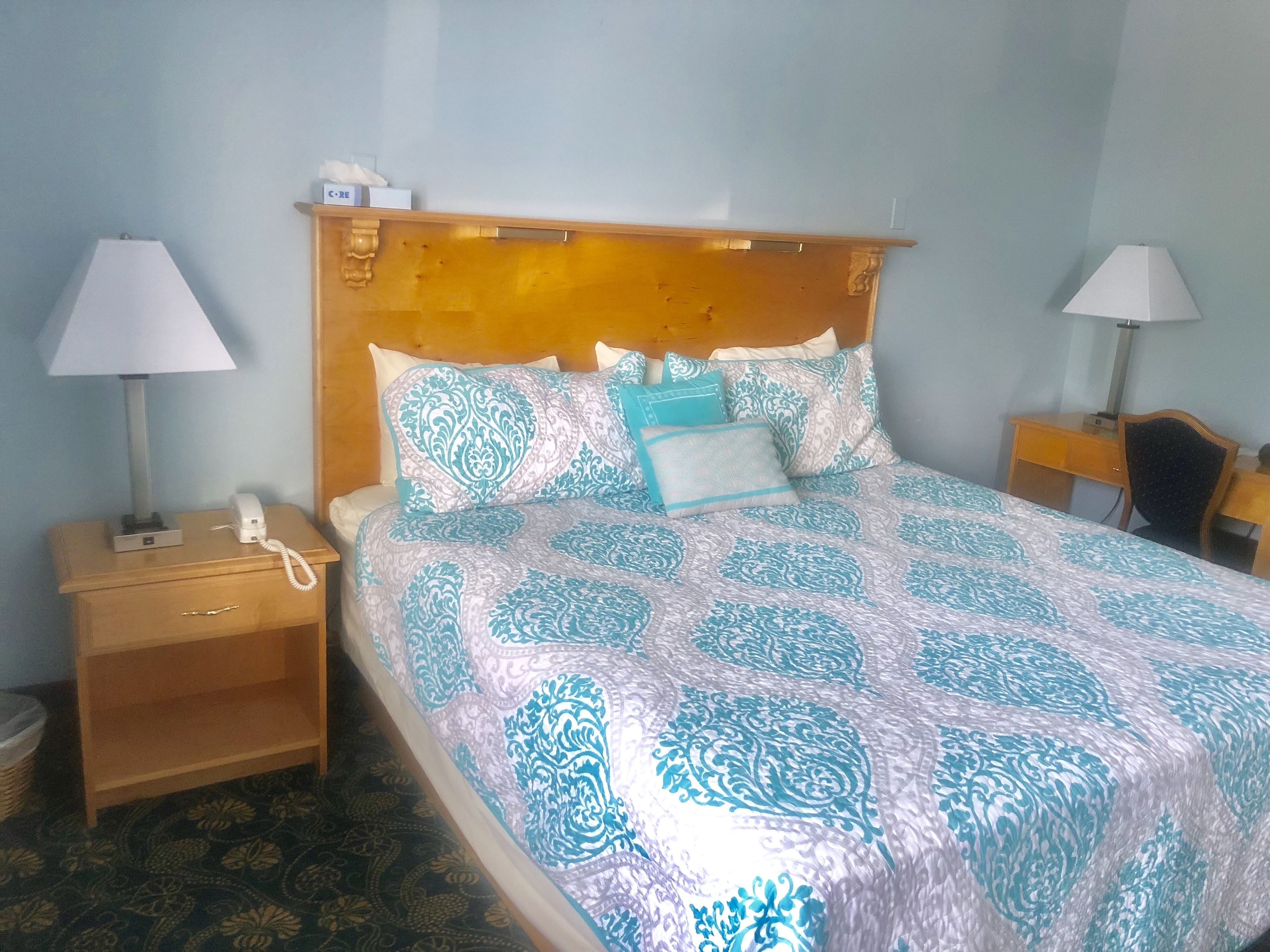 Uma cama bem arrumada com roupa de cama estampada, um abajur, um criado-mudo de madeira e uma pequena mesa com luminária.