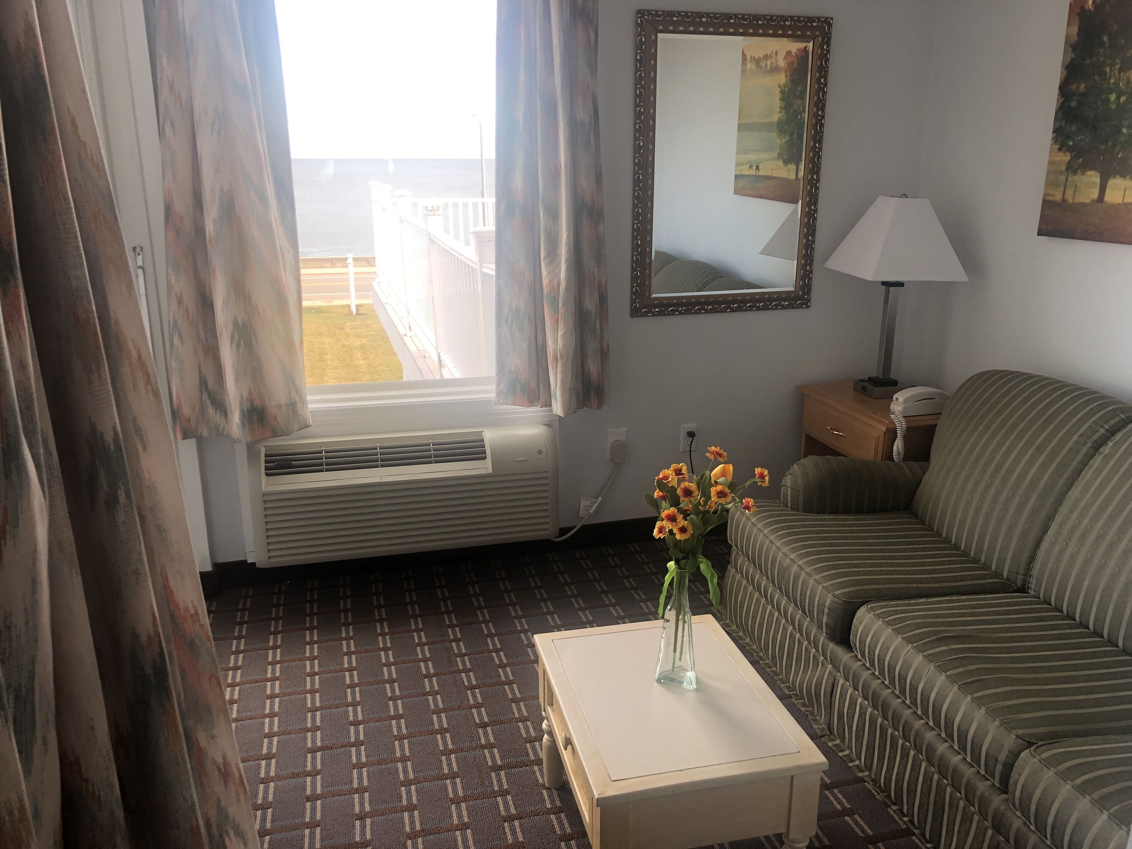A living room with a window, air conditioning unit, sofa, lamp, and a vase with flowers on a table.