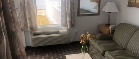 A living room with a window, air conditioning unit, sofa, lamp, and a vase with flowers on a table.