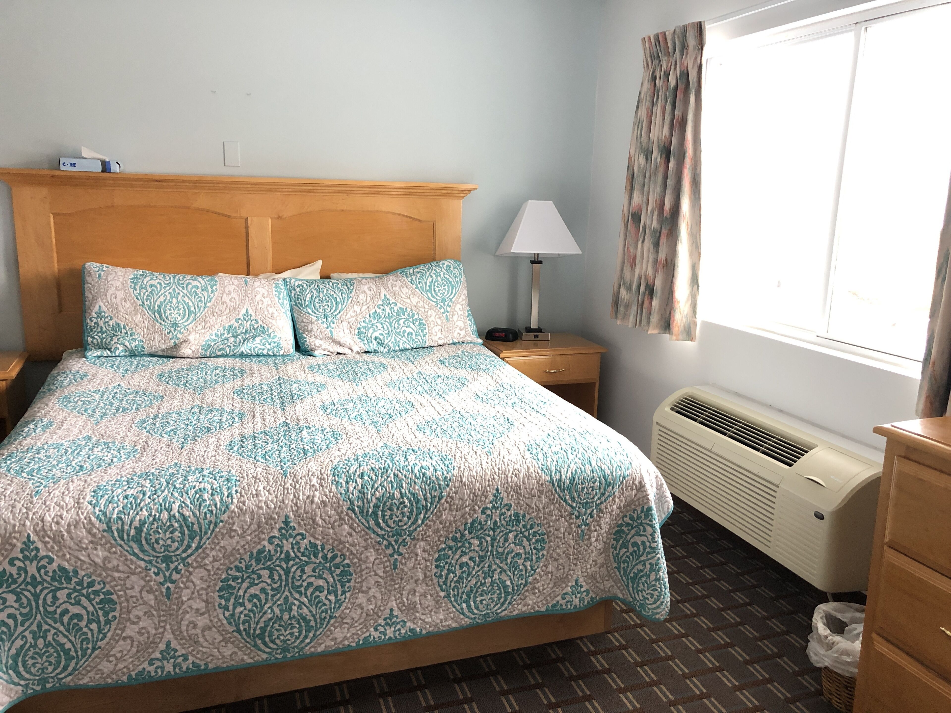 A neatly made bed with patterned bedding, a bedside lamp, a nightstand, a window with curtains, and a wall-mounted air conditioner.