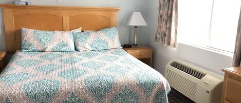 A neatly made bed with patterned bedding, a bedside lamp, a nightstand, a window with curtains, and a wall-mounted air conditioner.