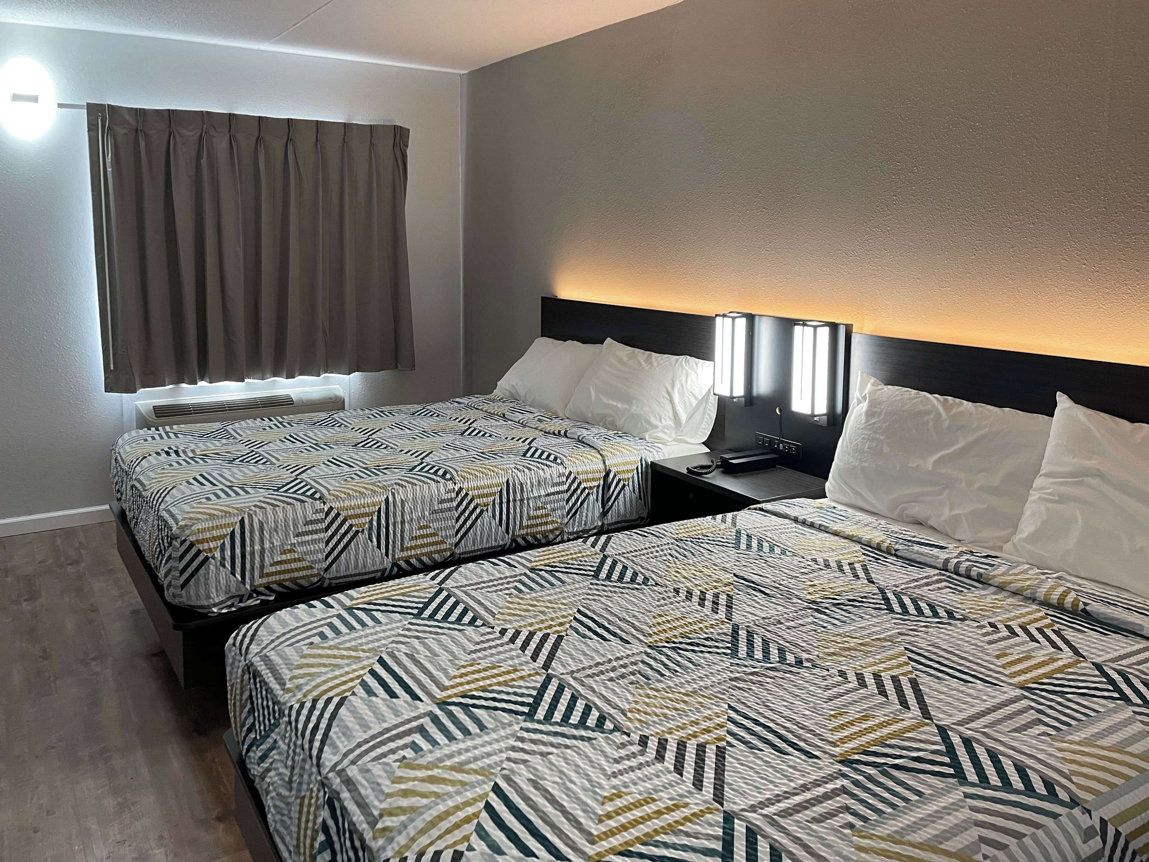 Habitación de hotel con dos camas, una ventana con cortinas, una mesita de noche con lámpara y un aplique de pared.