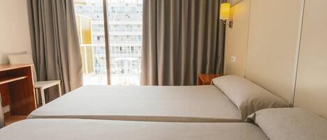 A hotel room with a large bed, a desk, a chair, a lamp, and a view of a building through the window.