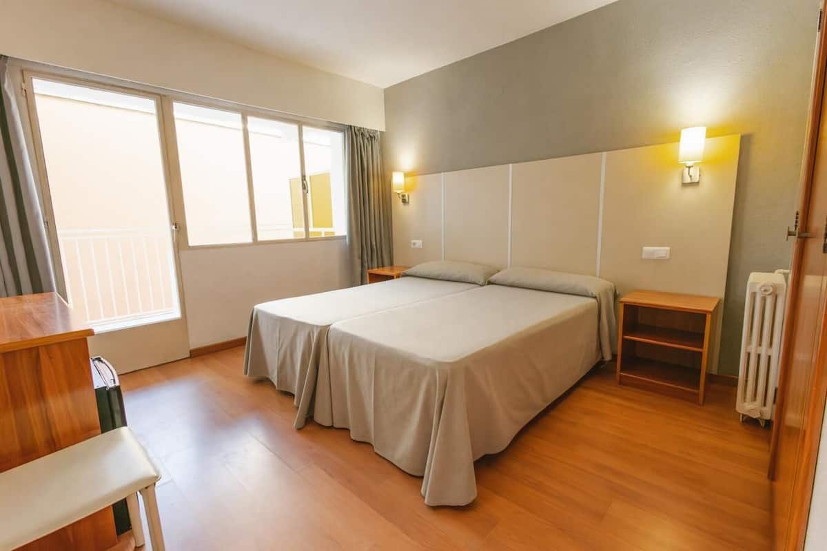 A hotel room with a double bed, a small table, a chair, a nightstand, a radiator, and a window with curtains.