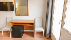 Une petite pièce avec un bureau en bois, deux chaises blanches, un meuble de rangement noir, un téléviseur fixé au mur et une fenêtre avec des rideaux.