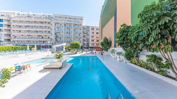 Uma piscina com água azul cristalina, rodeada por pavimento de azulejos brancos, e adjacente a um edifício com vários andares e varandas.