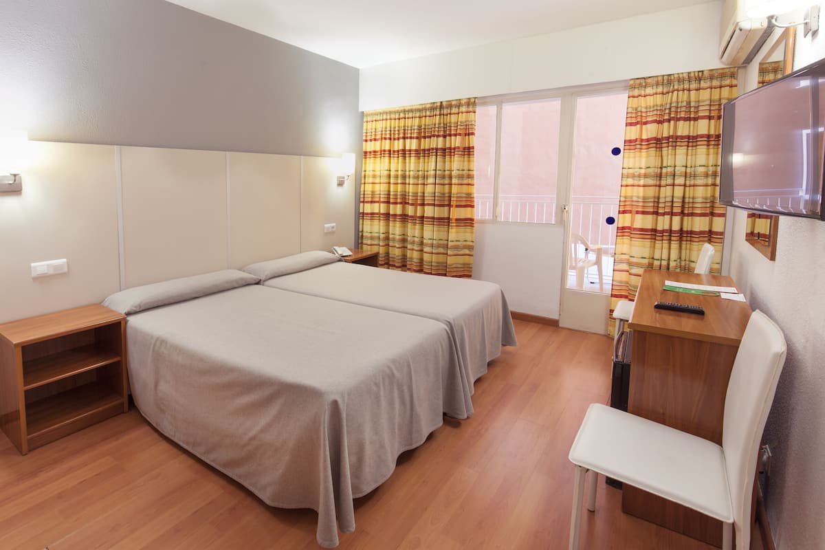 A hotel room with two beds, a desk with a chair, a TV, and a balcony with curtains.