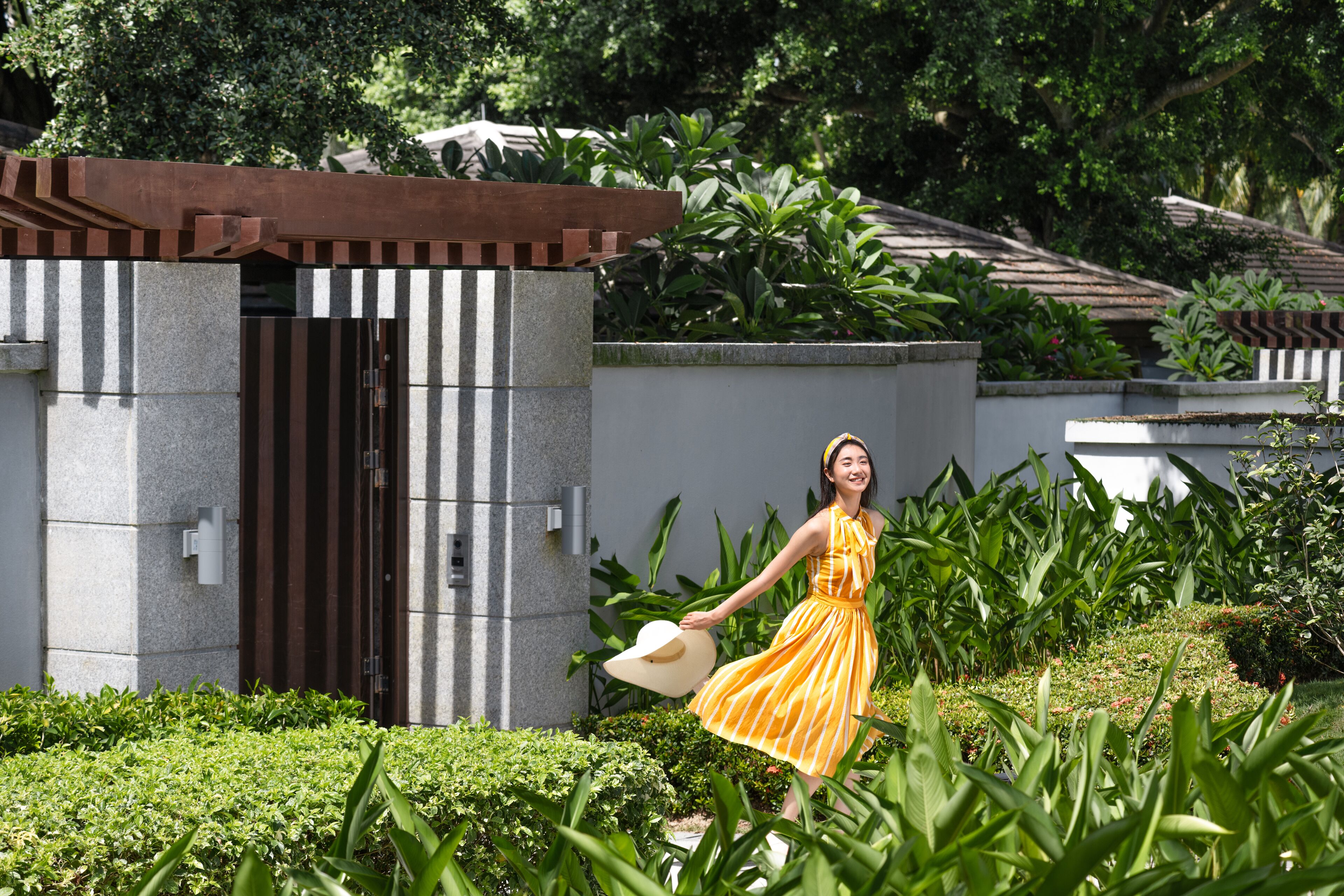 A woman in a yellow dress walking in a garden with a building in the background.