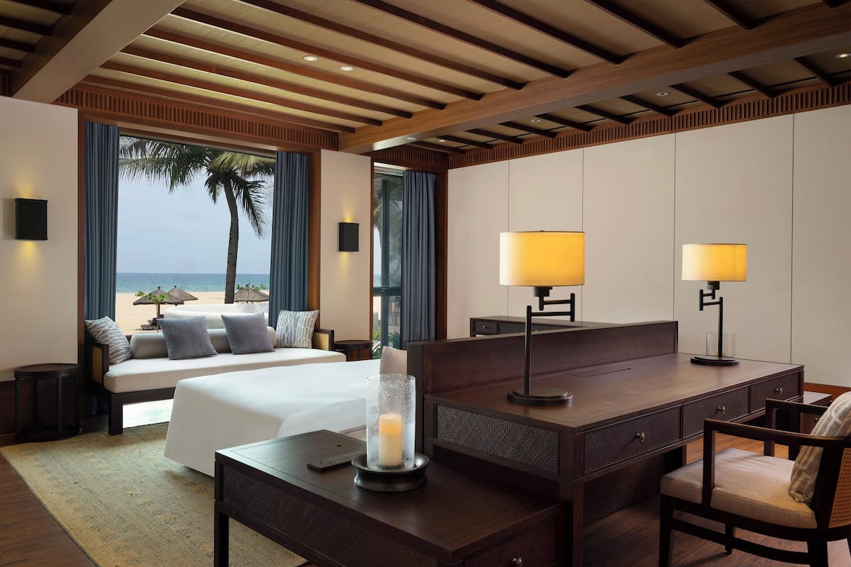 Une chambre d’hôtel avec un lit, un bureau et une chaise, une grande fenêtre donnant sur la plage et une table de chevet avec une lampe.