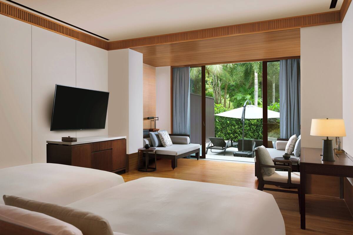 Une chambre d’hôtel dotée d’un grand lit, d’une télévision, d’un bureau avec une lampe, d’un coin salon avec une table et des chaises, et offrant une vue sur la verdure environnante.