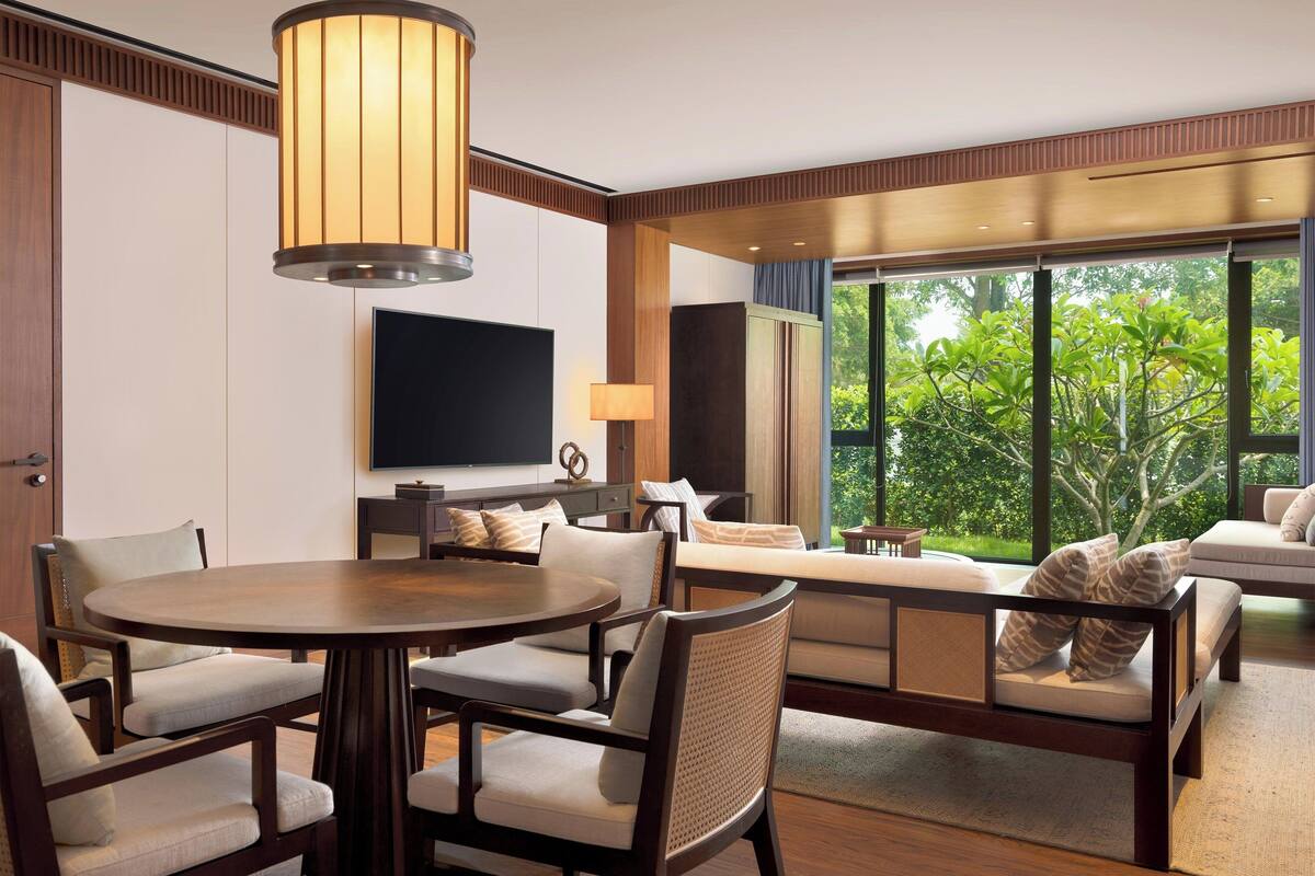 Une chambre d’hôtel moderne dotée d’une table de salle à manger ronde, de sièges confortables, d’un téléviseur à écran plat et d’une grande fenêtre offrant une vue sur la verdure.