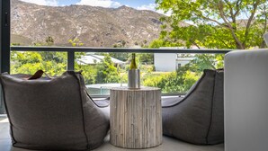 A balcony with two grey bean bag chairs, a small wooden table with a bottle and glasses, and a view of mountains and greenery. - Mimosa Lodge (Montagu)