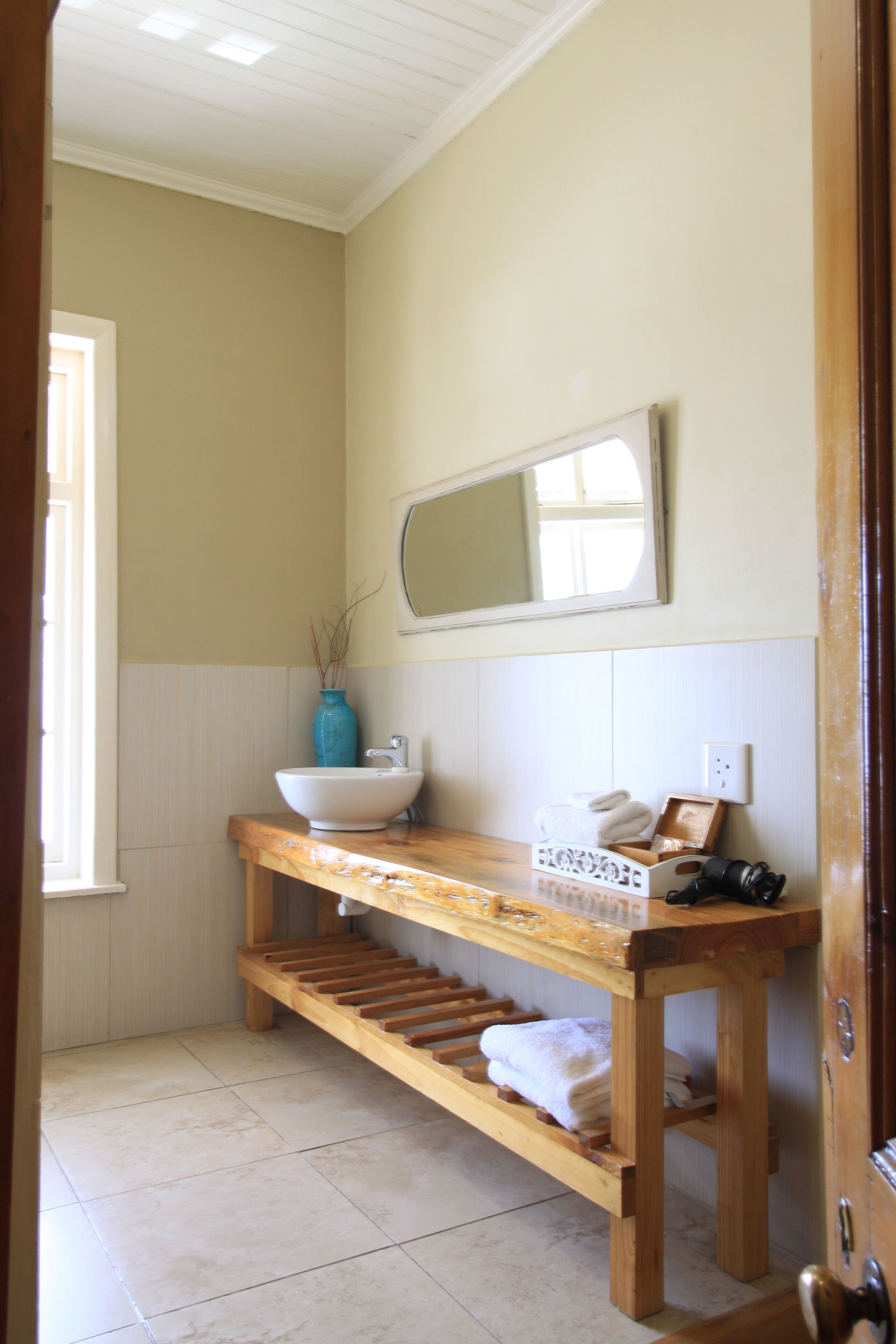 Un baño con un banco de madera, un lavamanos blanco, un florero azul y un espejo en la pared.