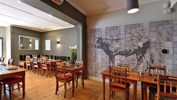 A restaurant with wooden tables and chairs, a large wall mural of a cow, and a modern ceiling design.
