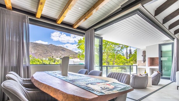A modern dining area with a wooden table and chairs, large windows with a mountain view, and a glass door leading to a balcony. - Mimosa Lodge (Montagu)