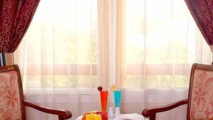 A dining area with a table set for two, a window with sheer curtains, and a view of greenery outside. - Safeer Hotel Suites - Aparthotel (Muscat)
