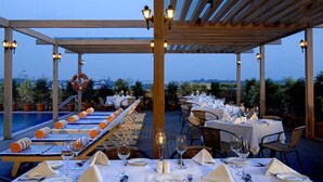 Un área de comedor al aire libre con mesas dispuestas para una comida, con vistas a una piscina y al paisaje urbano.