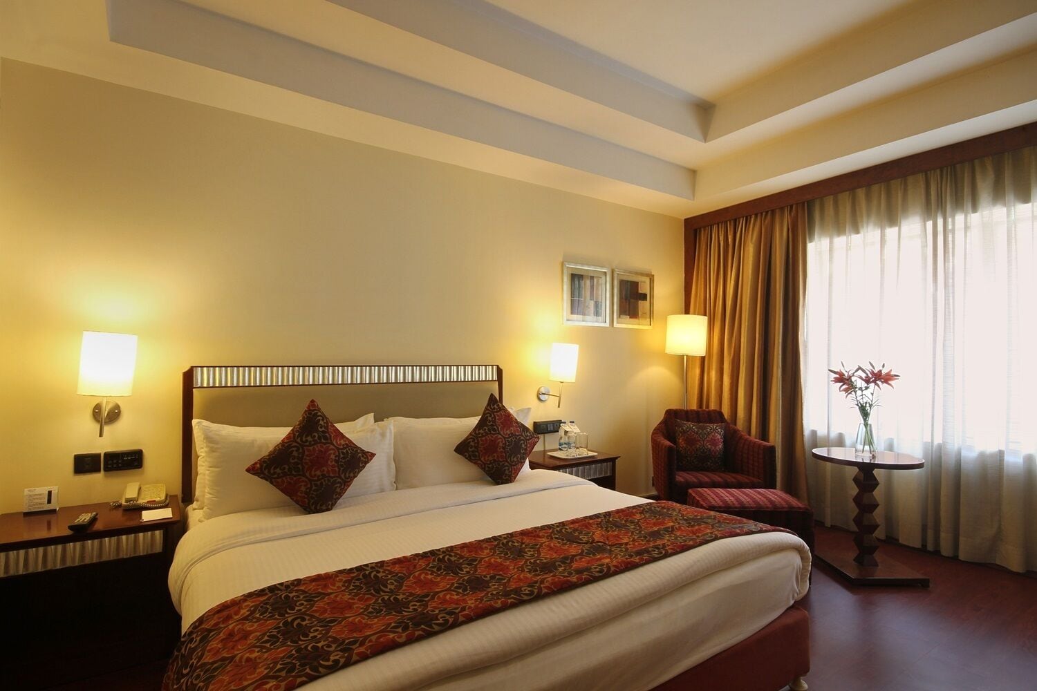 A hotel room with a large bed, two bedside lamps, a chair, a small table with a vase, and a window with curtains.