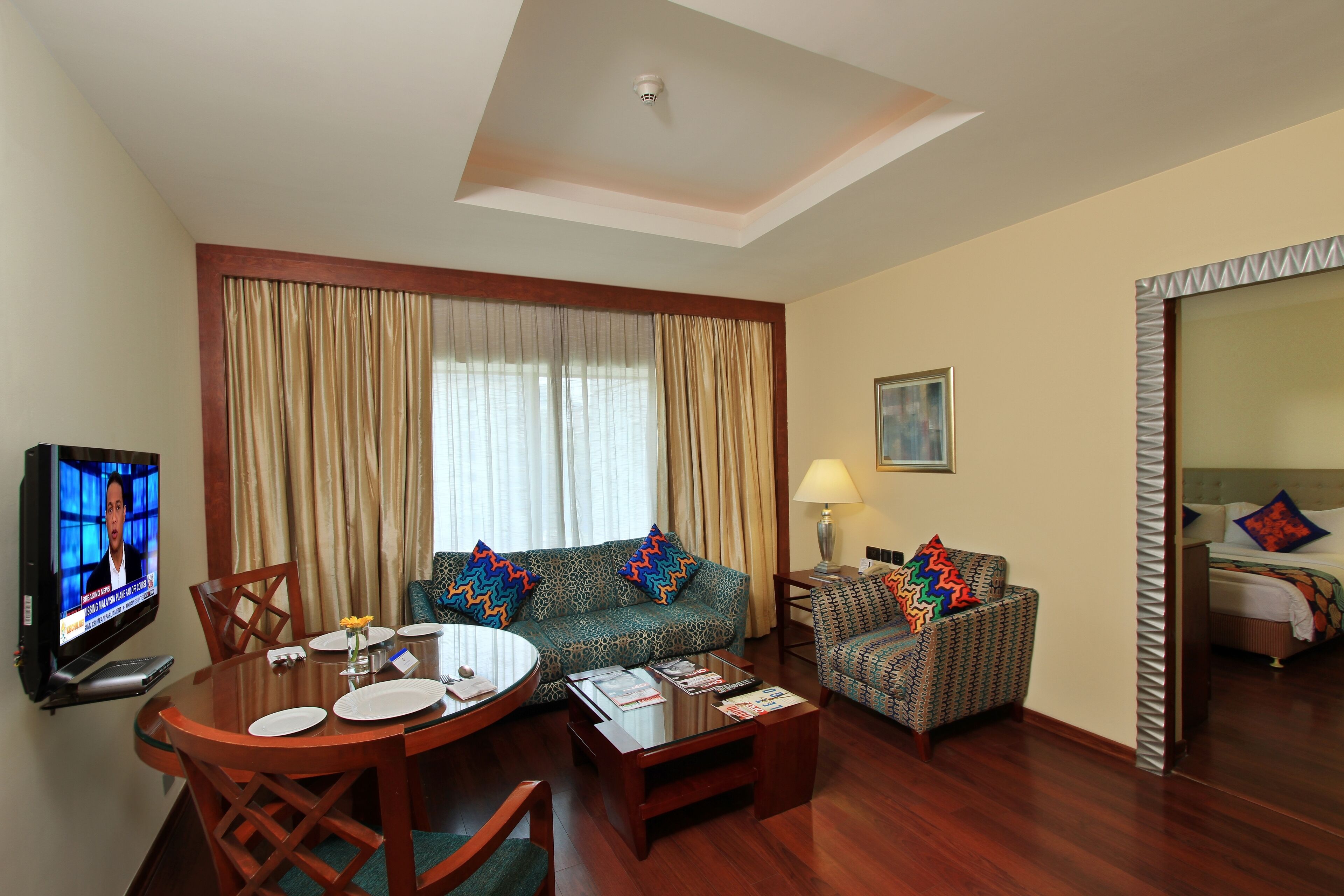 A hotel room with a dining area, a sofa, a patterned armchair, a TV, and a bed with a headboard.