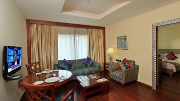 A hotel room with a dining area, a sofa, a patterned armchair, a TV, and a bed with a headboard.