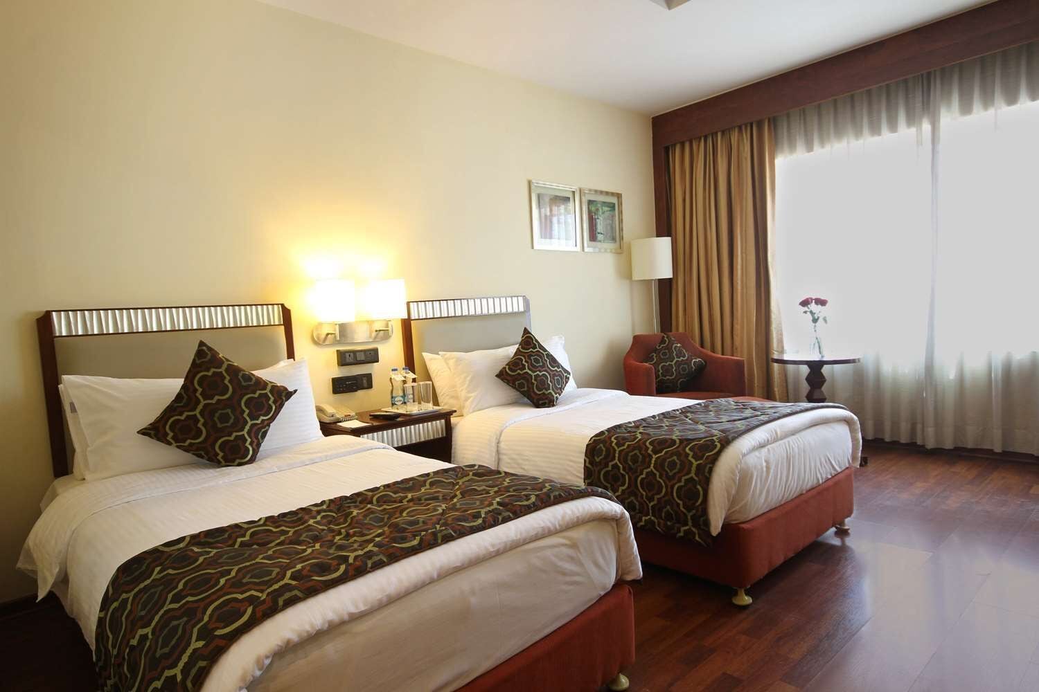 A hotel room with two beds, a nightstand, a lamp, a chair, a small table with a vase, and a large window with curtains.