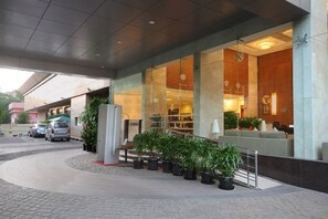 Una entrada de hotel moderna con fachada de vidrio, plantas de interior y un área de descanso con sofá y lámpara.