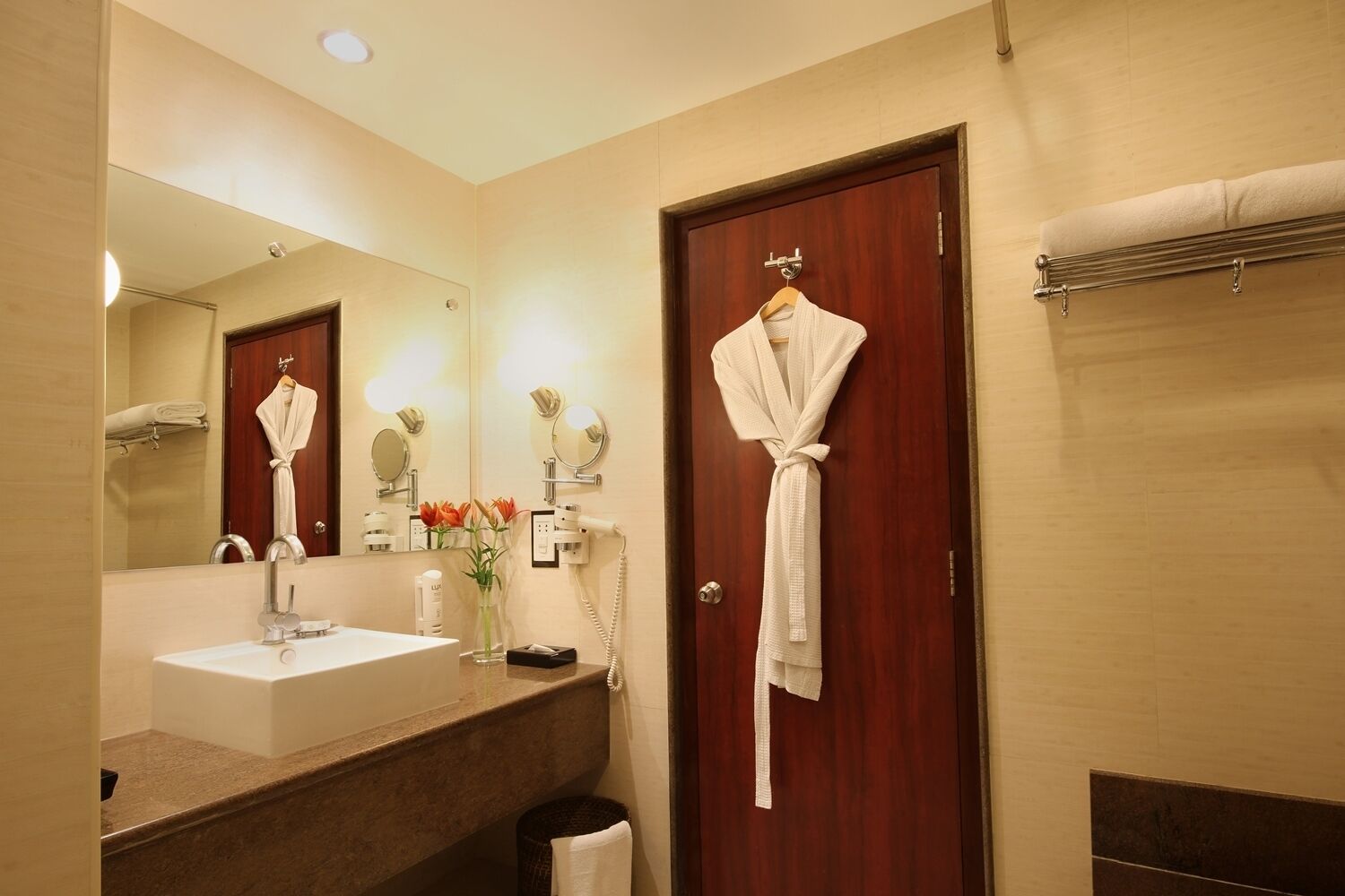 Un baño de hotel con un espejo grande, un lavamanos, una bata colgada en la puerta y un perchero para toallas.