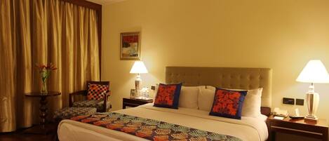 Habitación de hotel con una cama grande, dos mesitas de noche con lámparas, una silla, una pequeña mesa con un florero y un cuadro en la pared.
