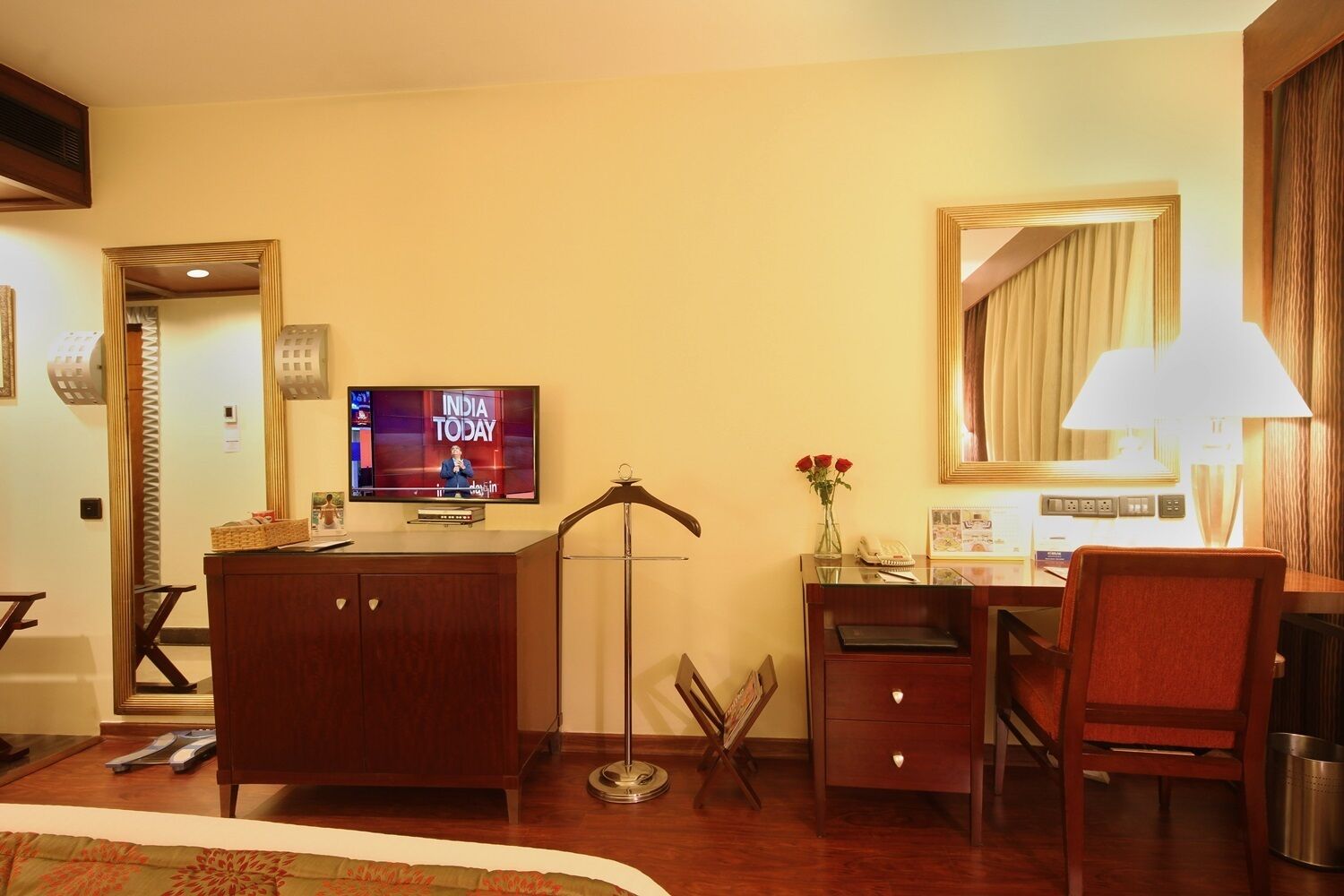 A hotel room with a wooden desk, a television displaying 'INDIA TODAY', a mirror, a lamp, and a vase with red flowers.