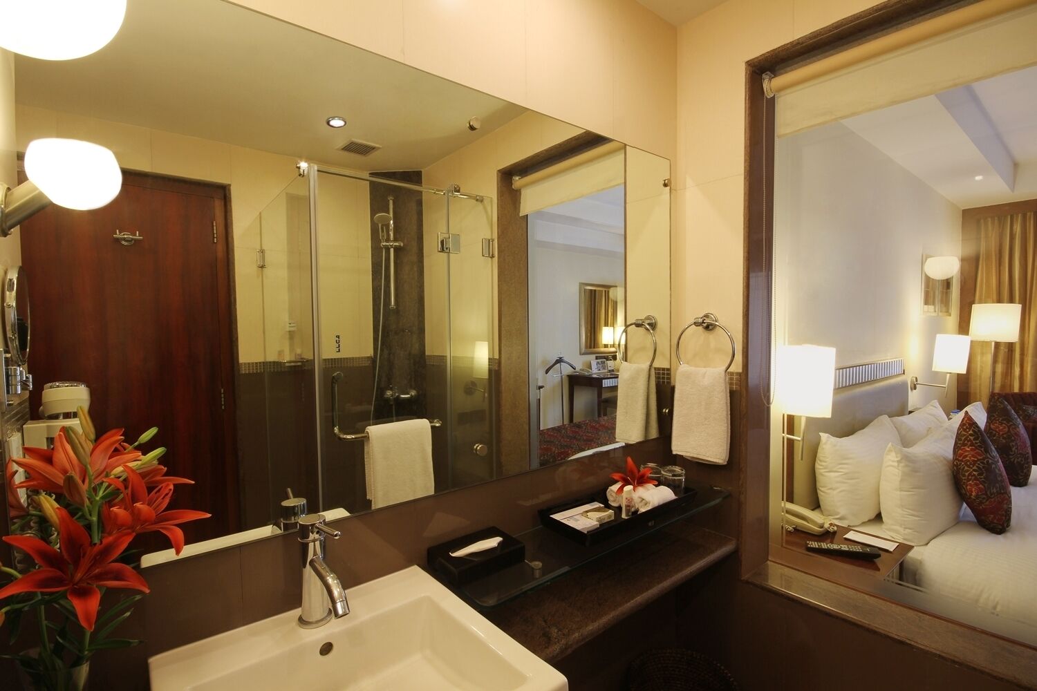 A hotel bathroom with a large mirror, a sink, a shower, and a bed with pillows and a blanket.