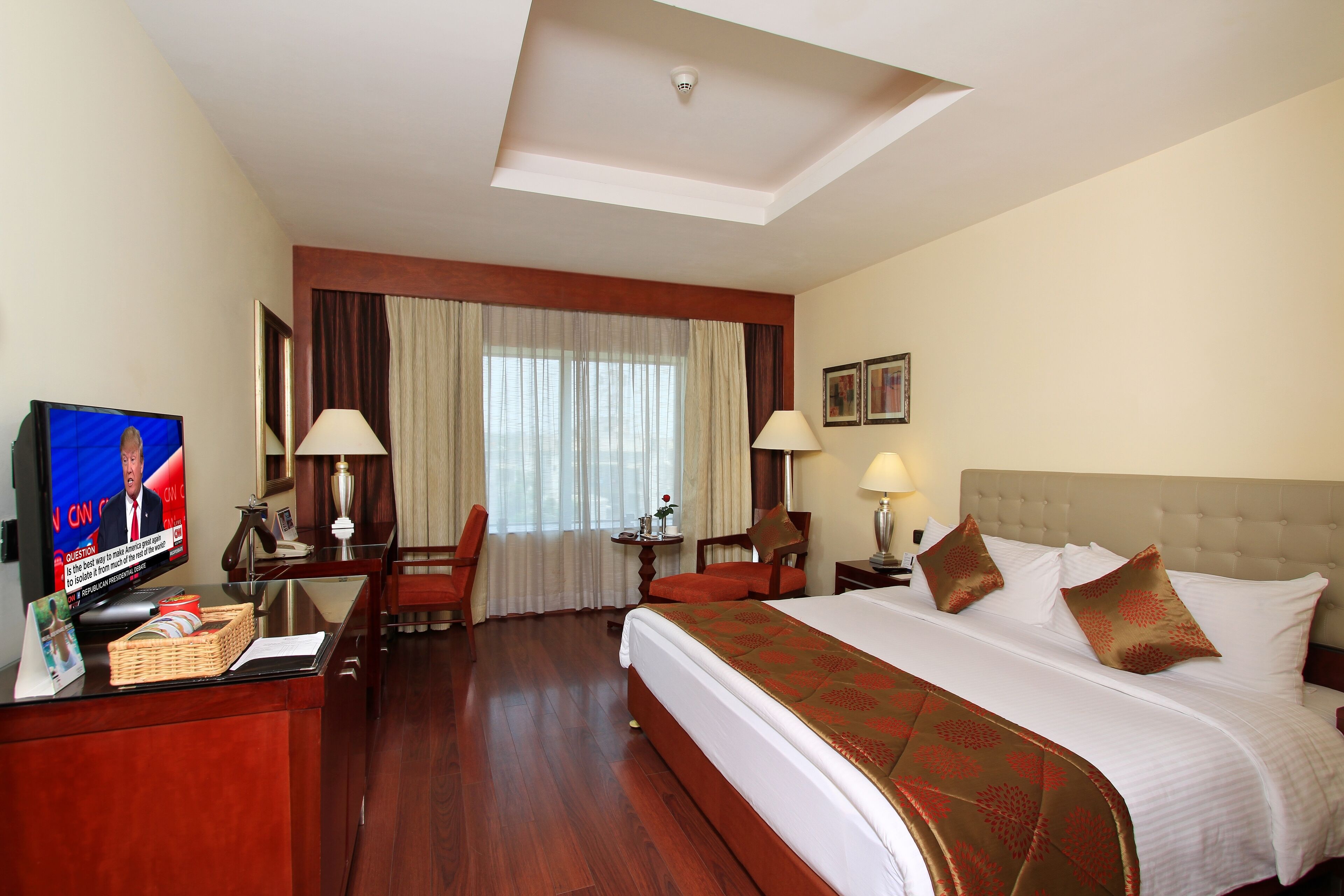 A hotel room with a large bed, a desk with a television, a chair, and a lamp.