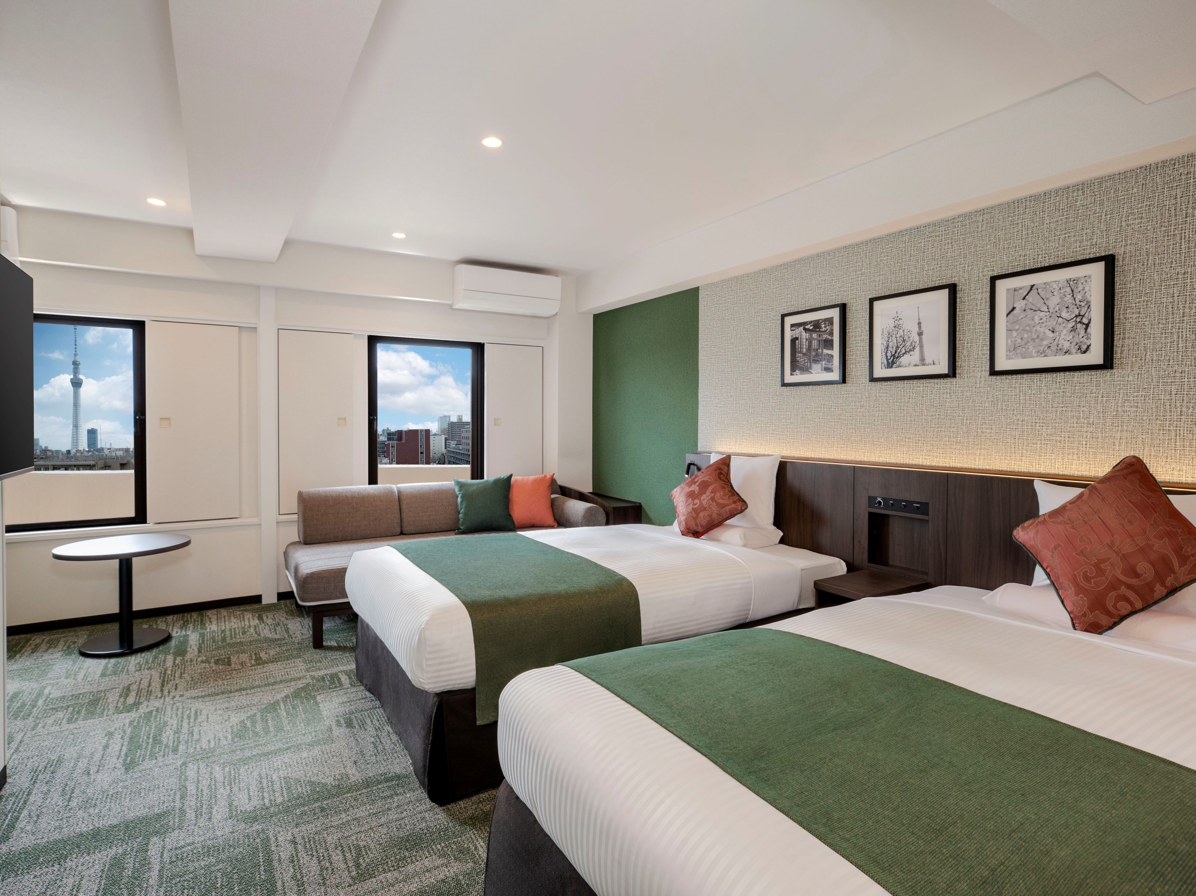 Habitación de hotel con dos camas, un sofá, una mesita y vista a un paisaje urbano a través del ventanal.