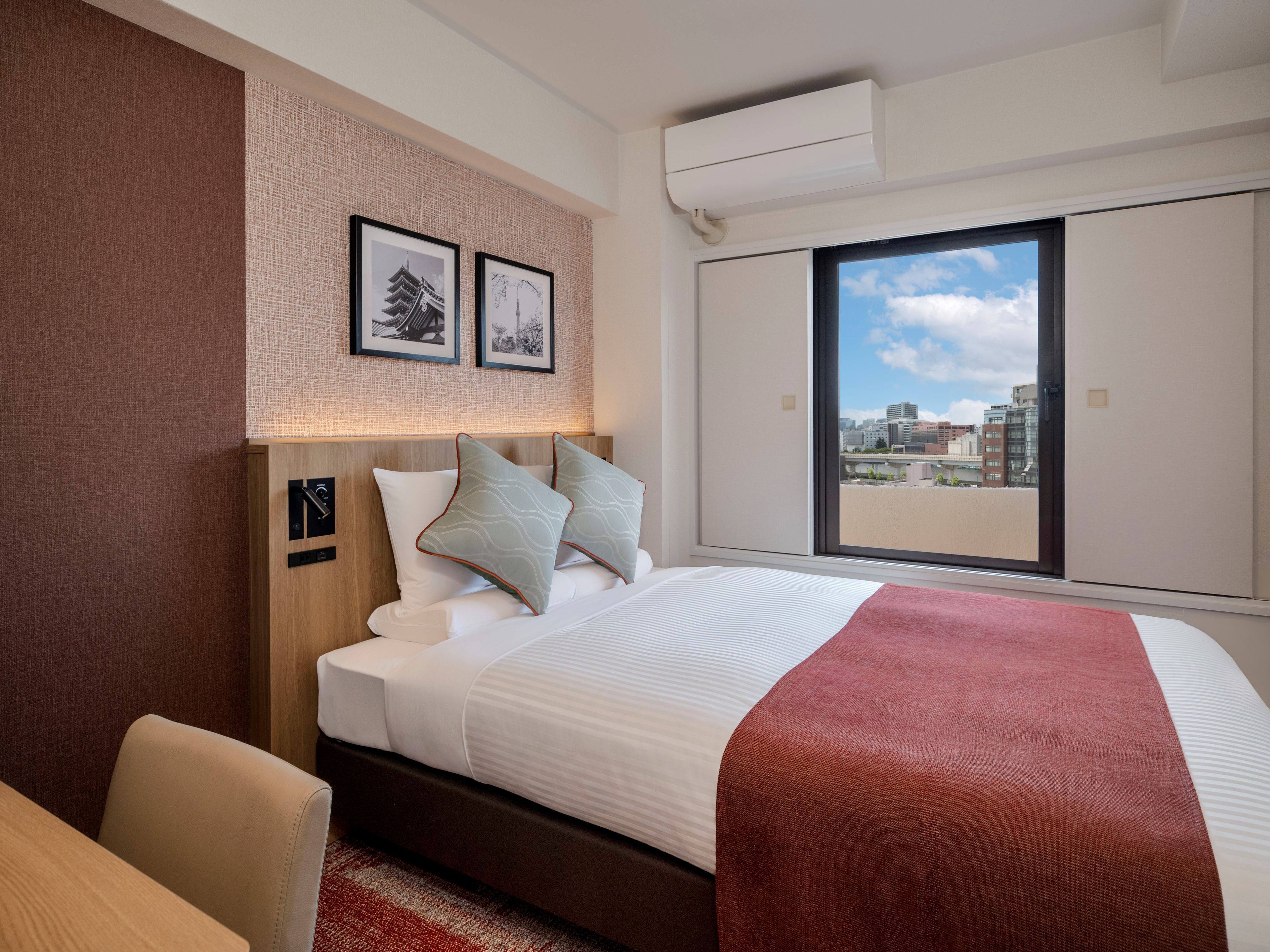 Ein Hotelzimmer mit Bett, Schreibtisch, Fenster mit Stadtblick und zwei gerahmten Bildern an der Wand.