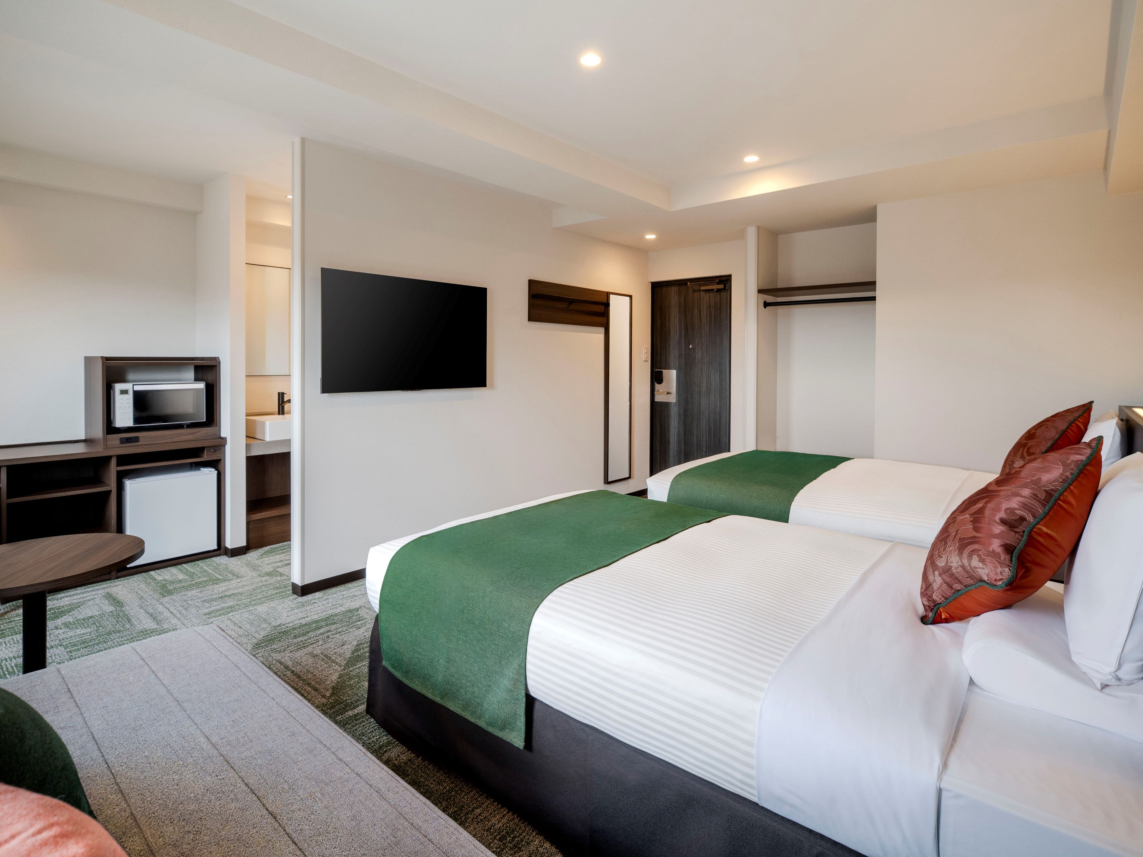 Ein Hotelzimmer mit zwei Betten, einem Schreibtisch mit Mikrowelle, einem Flachbildfernseher und einer Tür mit Spiegel.