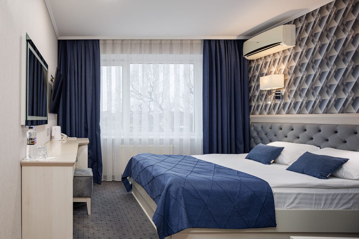 A hotel room with a bed, a desk, a chair, a mirror, and an air conditioning unit.