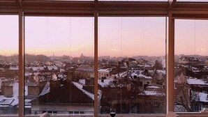 Una mesa de comedor con platos y una copa de vino, frente a un amplio ventanal con vistas a un paisaje urbano al atardecer.