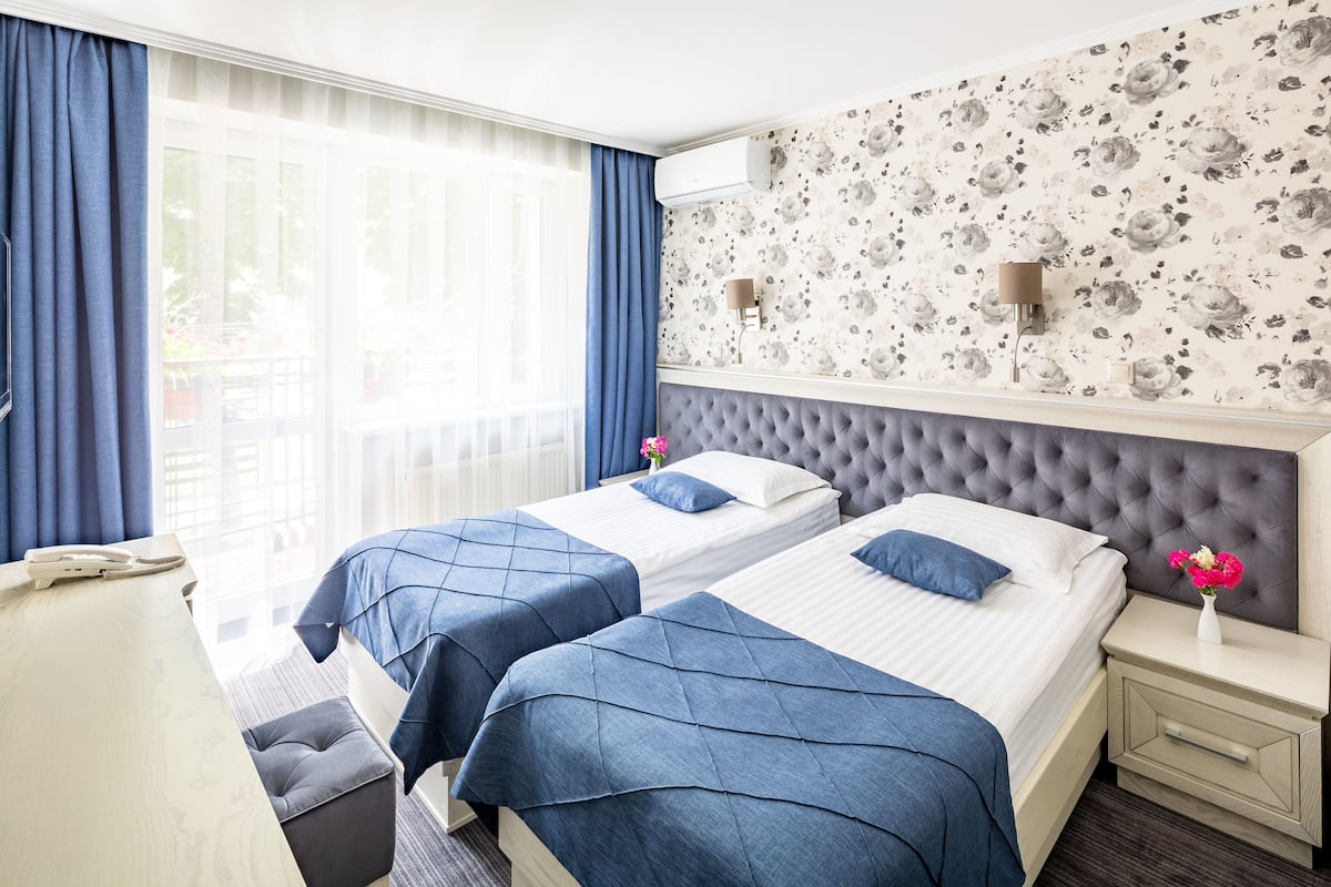 A hotel room with two beds, a large window with curtains, a wall-mounted air conditioner, and a small bedside table with a vase of flowers.