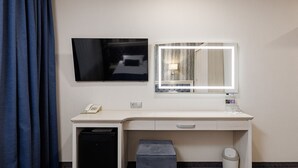 Habitación de hotel con escritorio, espejo, televisor, teléfono, cubo de basura y un taburete pequeño.