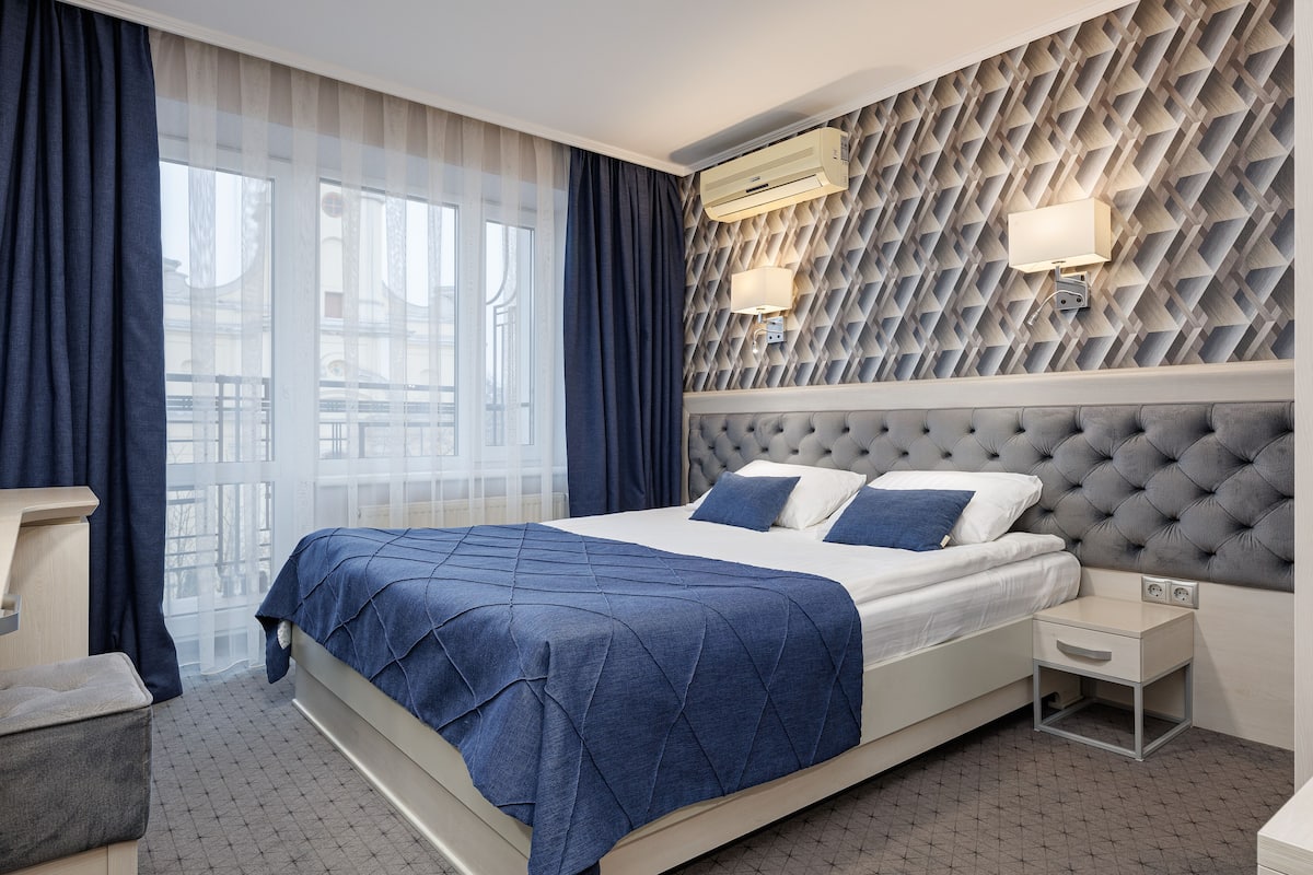 A modern hotel room with a large bed, a nightstand, a wall-mounted air conditioner, and a window with curtains.