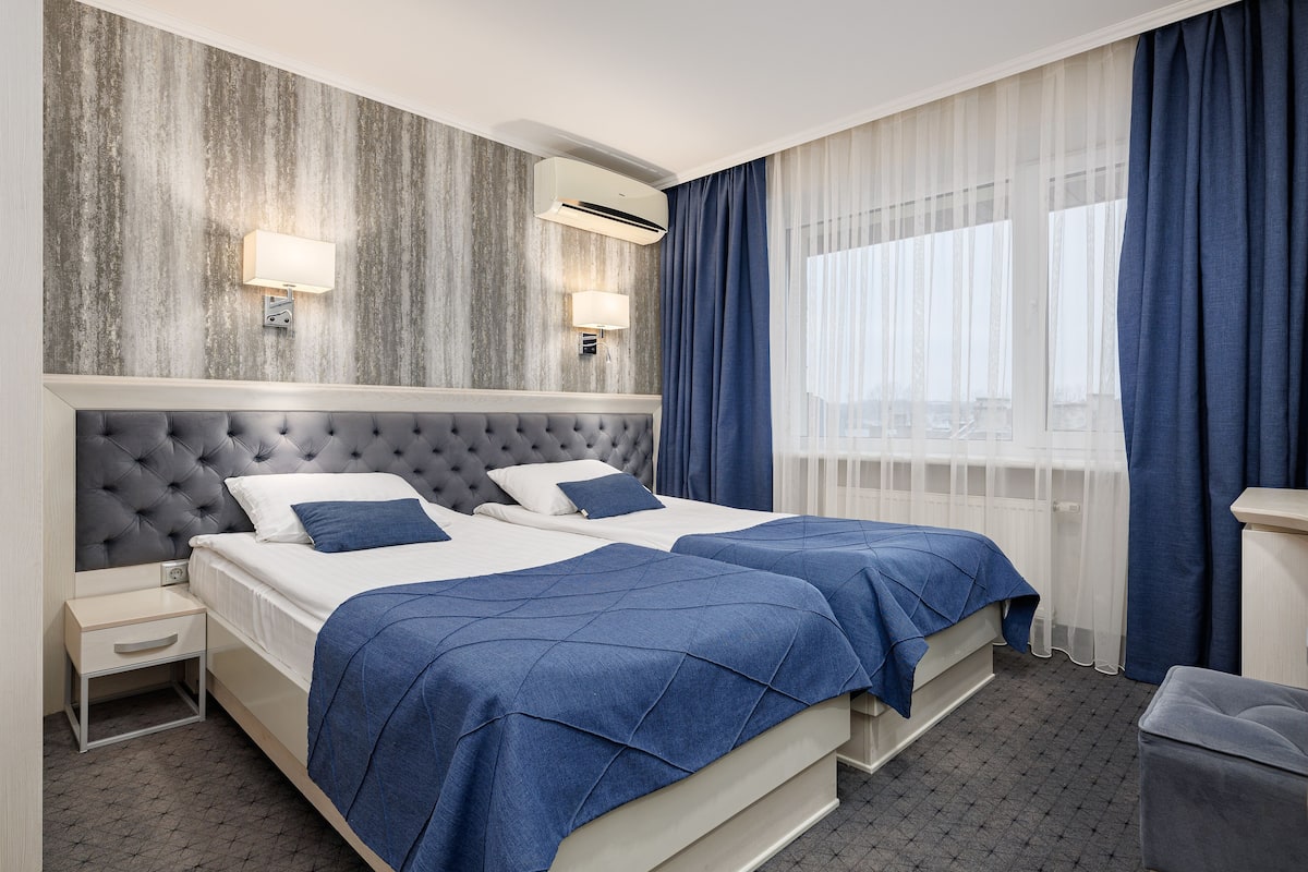 A hotel room with two beds, a grey tufted headboard, a white bedside table, a small desk, a trash bin, and a window with blue curtains.