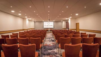Un salón de conferencias con hileras de sillas naranjas, piso alfombrado y una pantalla de proyección que muestra