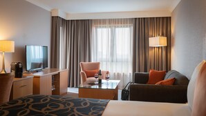 A hotel room with a bed, two armchairs, a coffee table, a TV, a desk with a lamp, and a window with curtains. - Clayton Hotel Leopardstown (Dublin)