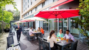 An outdoor dining area with tables and red umbrellas, surrounded by glass walls and a building with signage. - Clayton Hotel Leopardstown (Dublin)