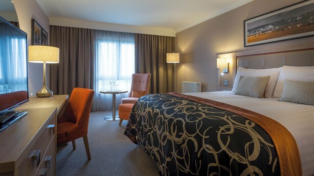 A hotel room with a bed, a desk, a chair, a table, and a window with curtains. - Clayton Hotel Leopardstown (Dublin)