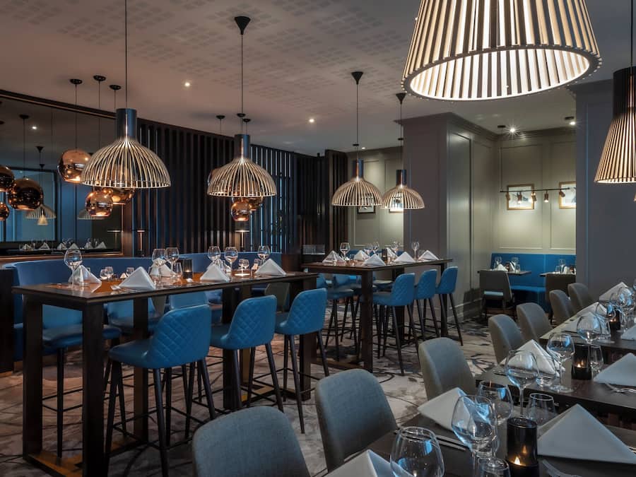 Een restaurant met meerdere eettafels, blauwe gestoffeerde stoelen en moderne hanglampen.