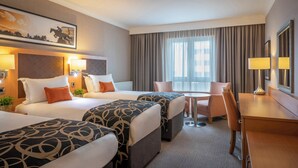 A hotel room with two beds, a desk, a chair, a small table, and a large window with curtains. - Clayton Hotel Leopardstown (Dublin)