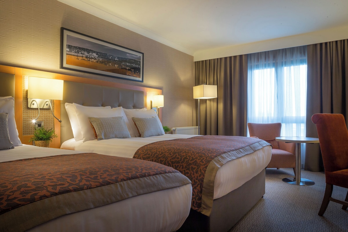 A hotel room with two beds, a small table, a chair, and a large window with curtains.